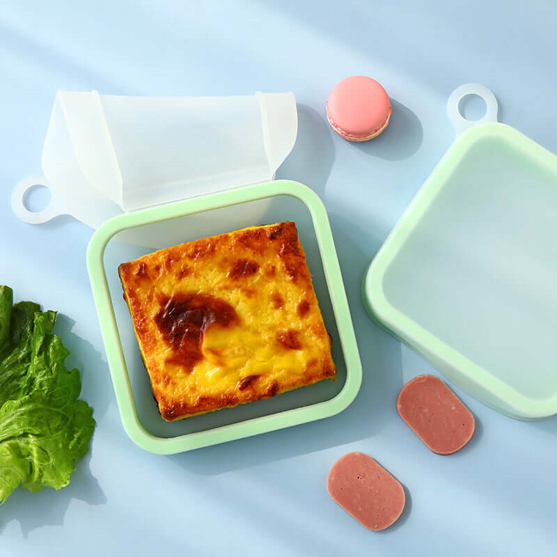 Square dish with cheesy food, surrounded by lettuce, pink condiment, and green lid on a light blue background